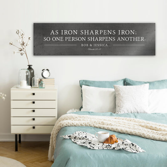 Metal sign with silver text that reads "As iron sharpens iron; so one person sharpens another." The sign also includes the names "Bob & Jessica" and the scripture reference "Proverbs 27:17".