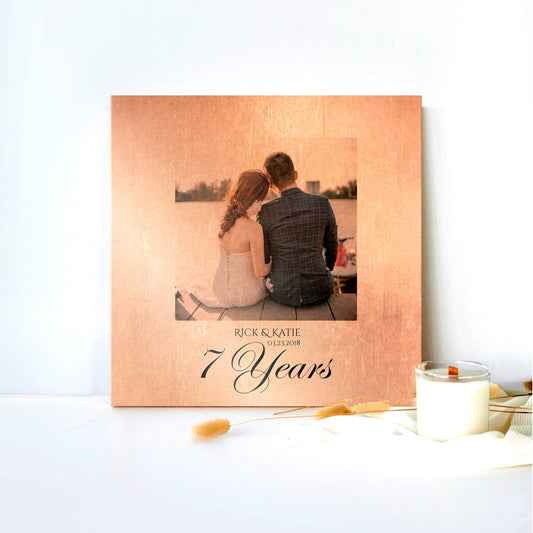Copper 7 Year Anniversary Photo Plaque is shown leaned against a white backdrop with a candle and silk cloth on the lower right of the frame. The copper plaque centers the photograph center in a square shape with the names of the couple in the photo beneath, and "7 Years" in a large calligraphy at the bottom. The plaque is distressed with rustic, vintage style with the photo blending with hues of the copper.
Courtesy of Honeycomb Proverbs