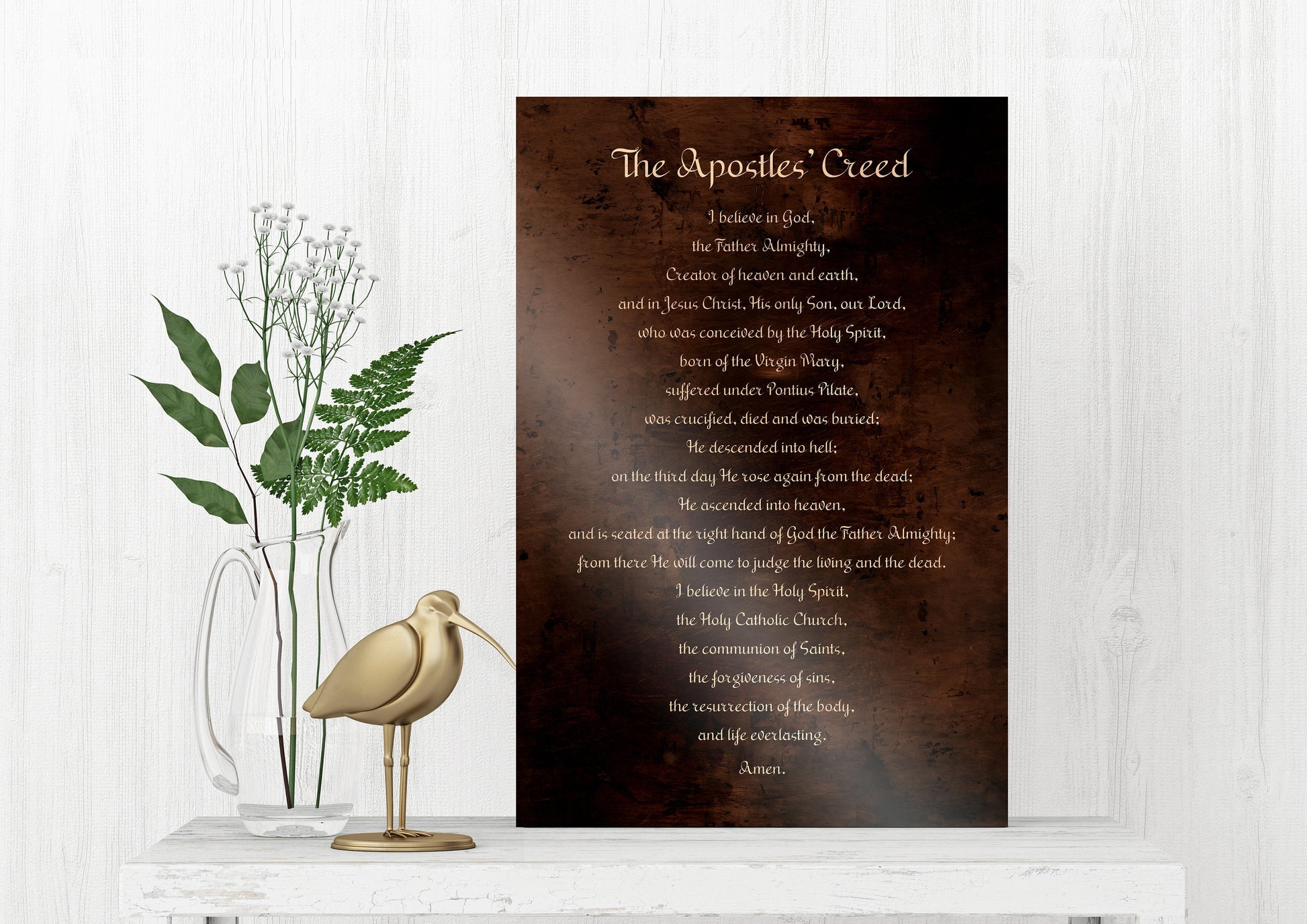 bronze, religious wall, art, typography plaque, or sign in rubbed bronze feature text from'The Apostles' Creed' in white lettering. The creed is written in light toned lettering on a bronze metal background. The sign is displayed on a white shelf with a wooden bird statue on it’s s left and greenery accents in a vase is set off to the left behind the bird statue.