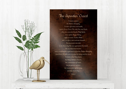 bronze, religious wall, art, typography plaque, or sign in rubbed bronze feature text from'The Apostles' Creed' in white lettering. The creed is written in light toned lettering on a bronze metal background. The sign is displayed on a white shelf with a wooden bird statue on it’s s left and greenery accents in a vase is set off to the left behind the bird statue.