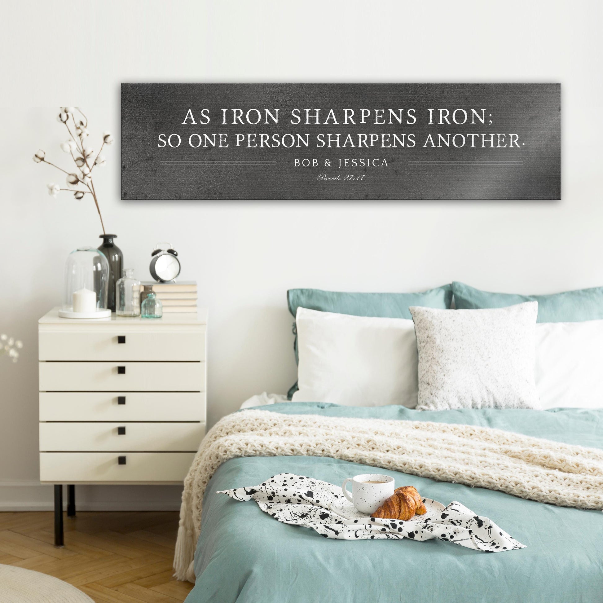 Metal sign with silver text that reads "As iron sharpens iron; so one person sharpens another." The sign also includes the names "Bob & Jessica" and the scripture reference "Proverbs 27:17".