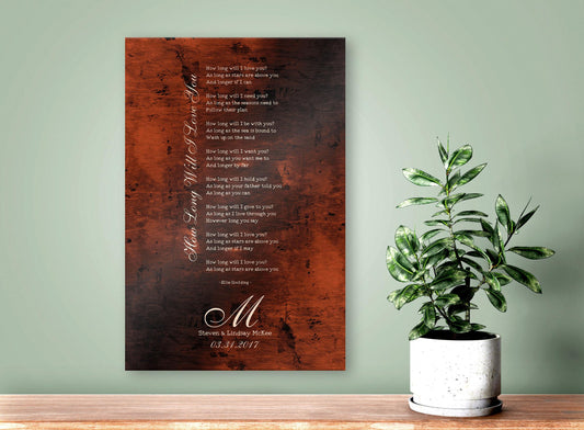 A burnished bronze anniversary gift plaque is personalized with a calligraphic monogram letter,  the wedding anniversary date and names of a married couple. Above is their special wedding song, laid out in a traditional lyrical stanza with the song title elegantly placed vertically, along the left margin. The wedding song lyric art is wall hung on a sage green wall above a shelf where a potted plant also accents the artwork on the right hand side of the frame.