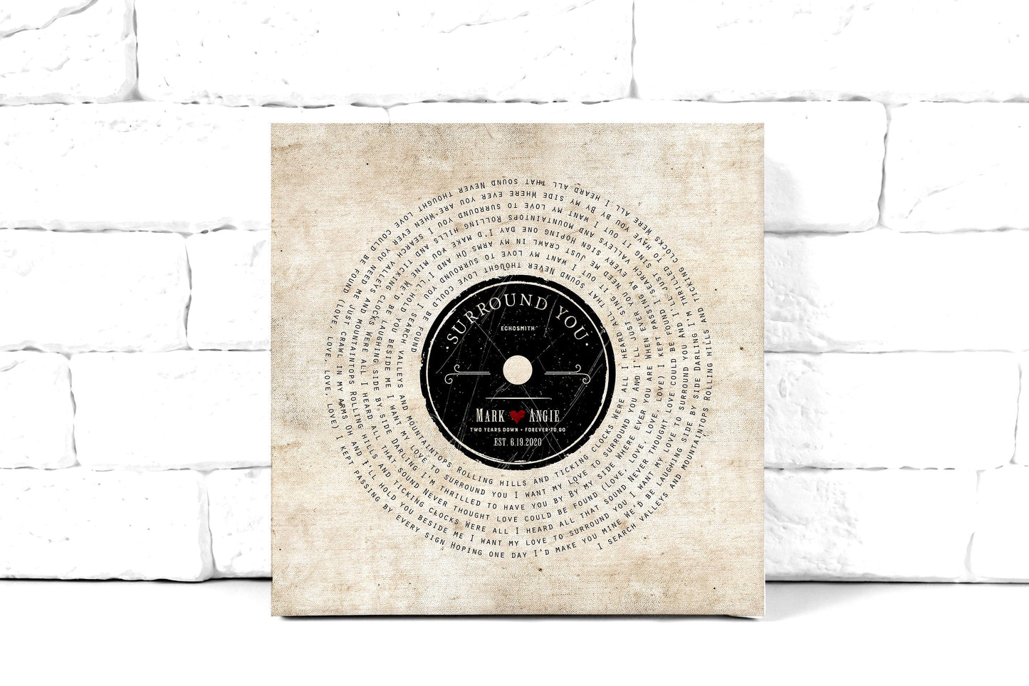 lyrics on canvas, custom song art with lyrics set in a vinyl record design around a retro record label personalized with a couple's first names, with a heart stamp, and the song name and performing artist of choice. Art also features the statement "2 years down, forever to go" as a nod to the 2nd wedding anniversary. This cotton canvas song art is leaning against a white brick wall.