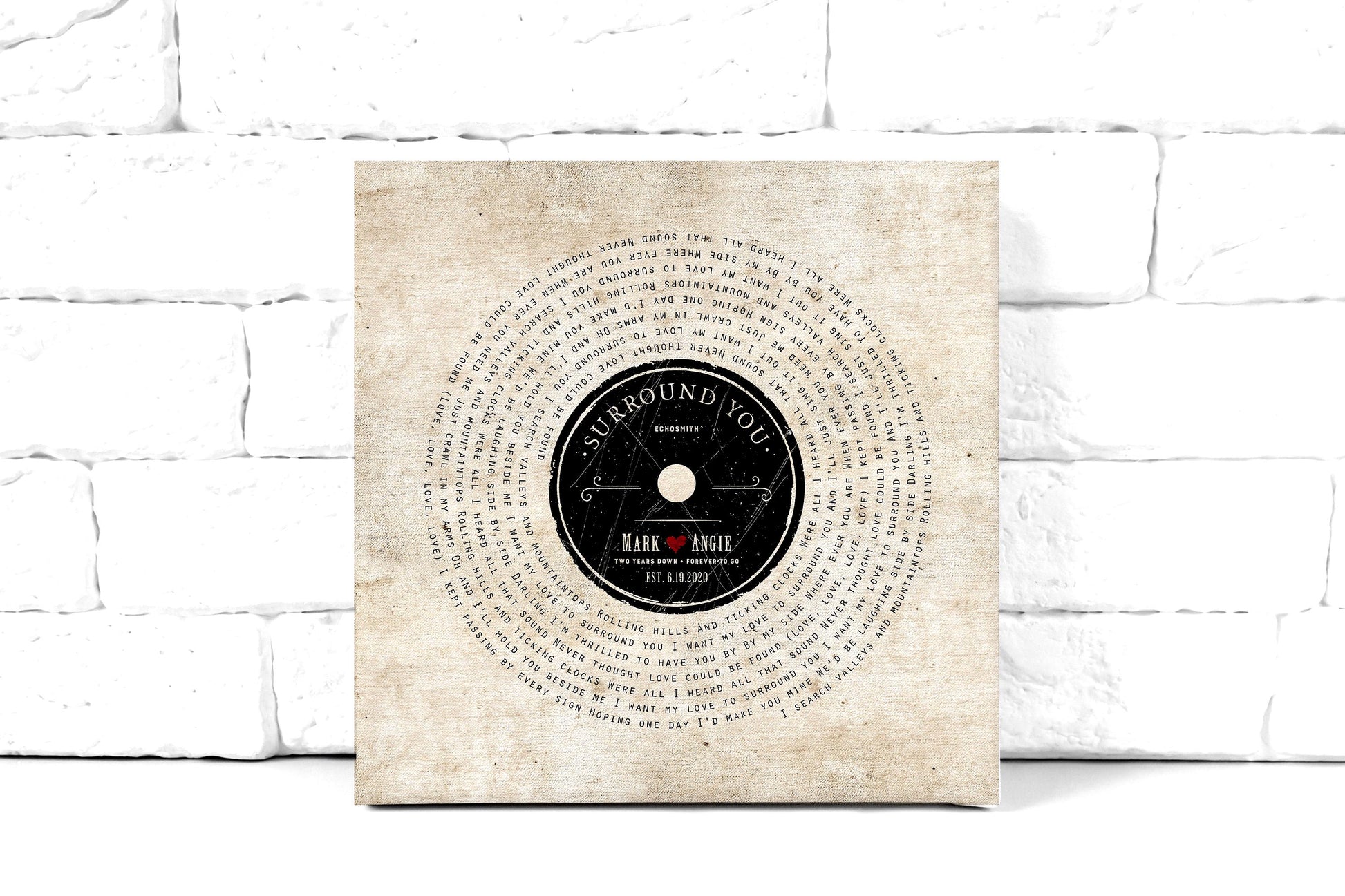 lyrics on canvas, custom song art with lyrics set in a vinyl record design around a retro record label personalized with a couple's first names, with a heart stamp, and the song name and performing artist of choice. Art also features the statement "2 years down, forever to go" as a nod to the 2nd wedding anniversary. This cotton canvas song art is leaning against a white brick wall.