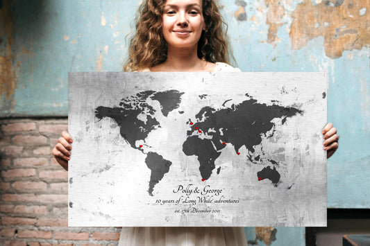 Woman holding large metal adventure map wall art. Wall art shown is a vintage tarnished and rustic world map on metal, personalized with names, an established date, and includes heart inscribed markers to "pin" locations that hold special meaning. This is a silver, grey and black metal sign with red toned hearts.

Photo courtesy of Honeycomb Proverbs
