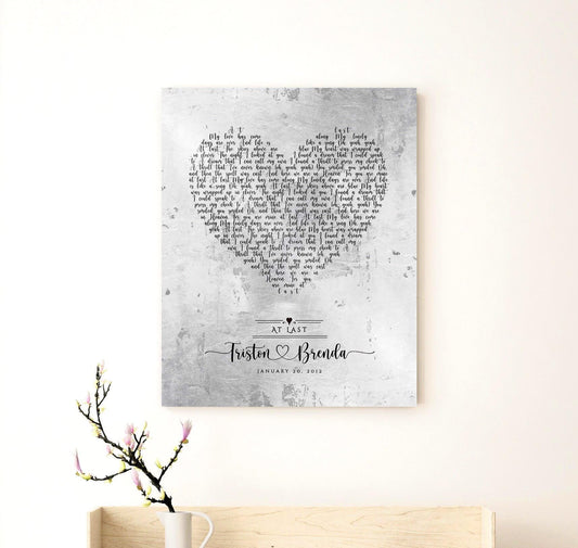 Metal Tin Song Art with Song Lyrics inscribed onto a distressed metal background and arranged in a heart shape. Personalized with couple's names connected by a heart and hung on a wall above a shelf with a vase and cut flowers.