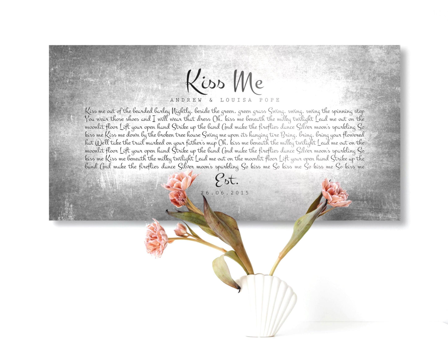 Tarnished Tin Anniversary Song Plaque hangs on wall above a vase with flowers. The tin sign is custom made and features the phrase "Kiss Me" and personalized song lyrics beneath, as requested by buyer. Also included are the names of the recipients and union/established date. The typography style that was selected for this custom piece is playful and romantic and starkly contrasts the metal sign.