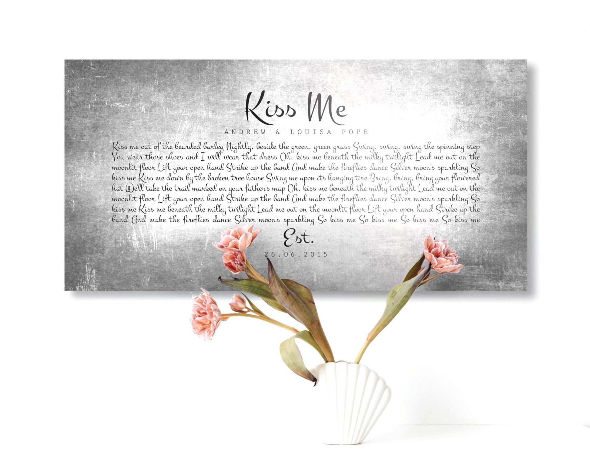 Tarnished Tin Anniversary Song Plaque hangs on wall above a vase with flowers. The tin sign is custom made and features the phrase "Kiss Me" and personalized song lyrics beneath, as requested by buyer. Also included are the names of the recipients and union/established date. The typography style that was selected for this custom piece is playful and romantic and starkly contrasts the metal sign.