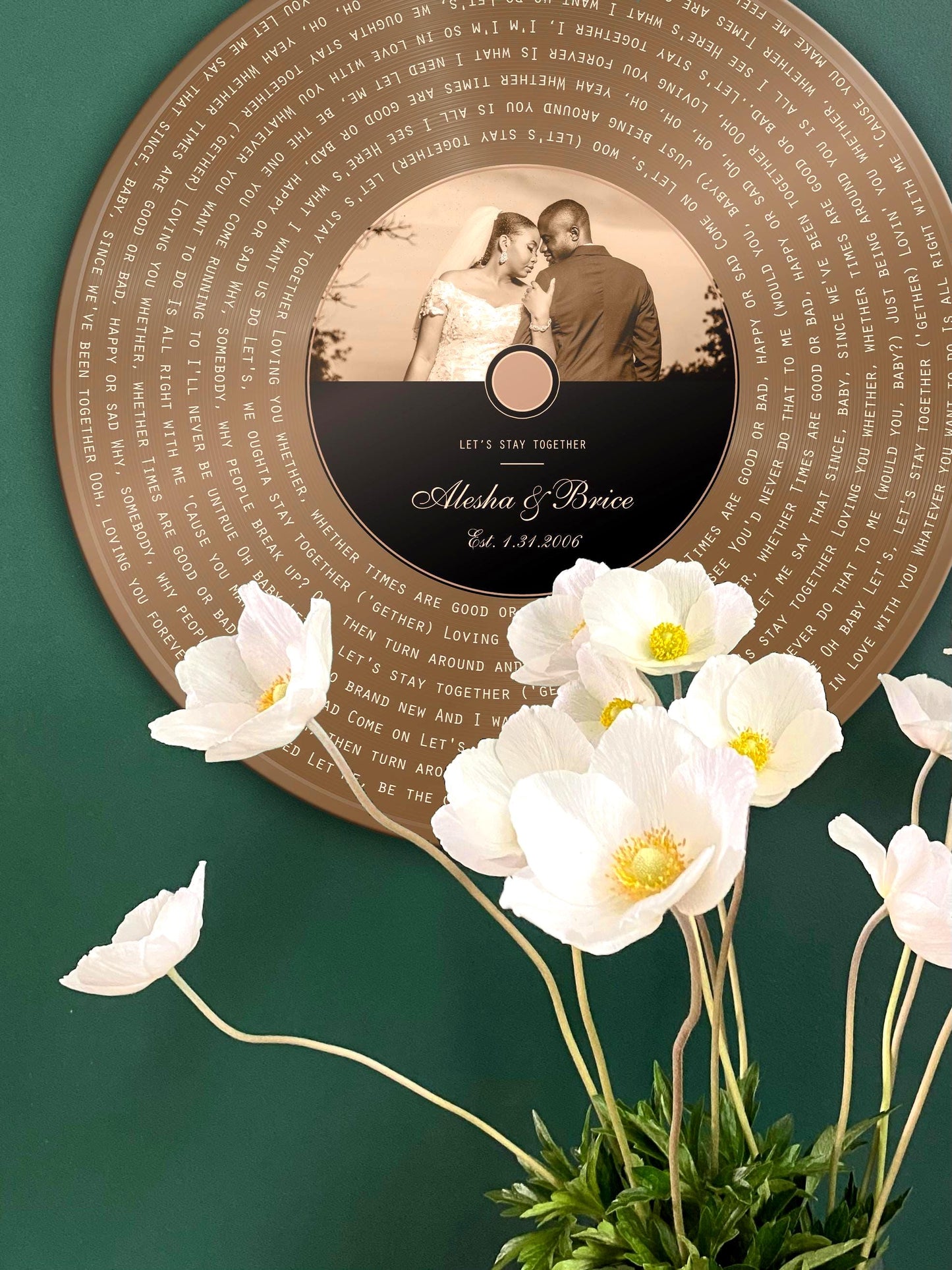 Personalized Bronze Anniversary record includes song lyrics of choice, overlaid onto the vinyl motif and photo and song credit with the couple's names are inscribed onto the record label area. Round Bronze metal record is hung on a green wall and white flowers delicately frame the lower third of the photo.  Photo courtesy of Honeycomb Proverbs