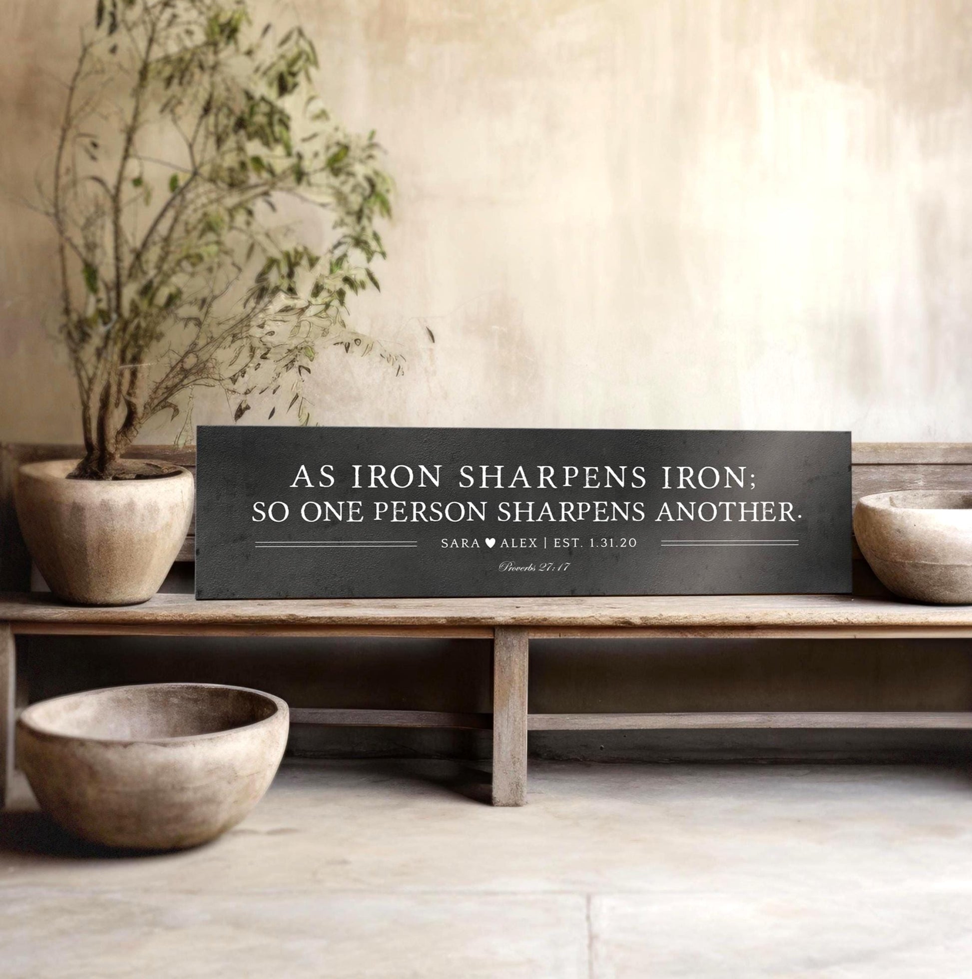 Metal sign with silver text that reads "As iron sharpens iron; so one person sharpens another." The sign also includes the names "Sara & Alex" and the scripture reference "Proverbs 27:17".