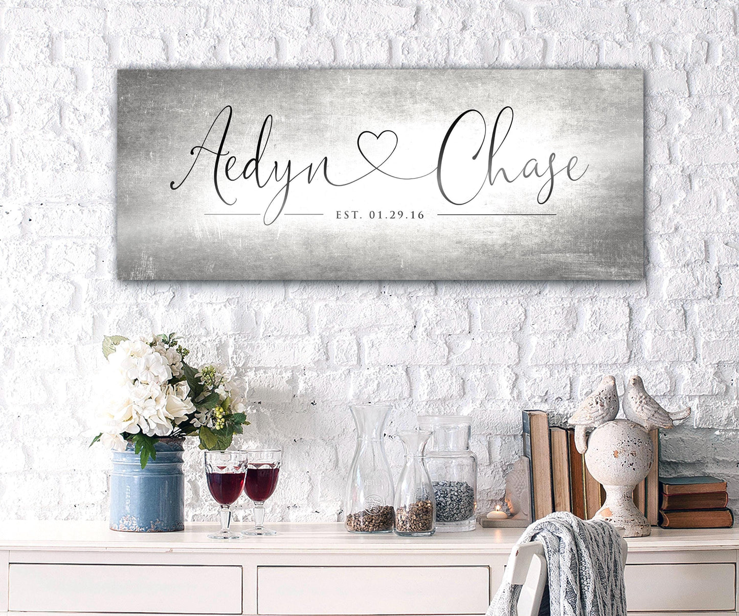 Unique Anniversary gifts like Honeycomb Proverbs' Personalized Heart Name signs are popular for ten year wedding anniversaries. One of their tarnished metal Heart Name sign hangs above an accent table. The sign is extra large sized and features personalized names connected by a heart in a whimsical calligraphy font. The couple's established date sits beneath their names to mark their special day.