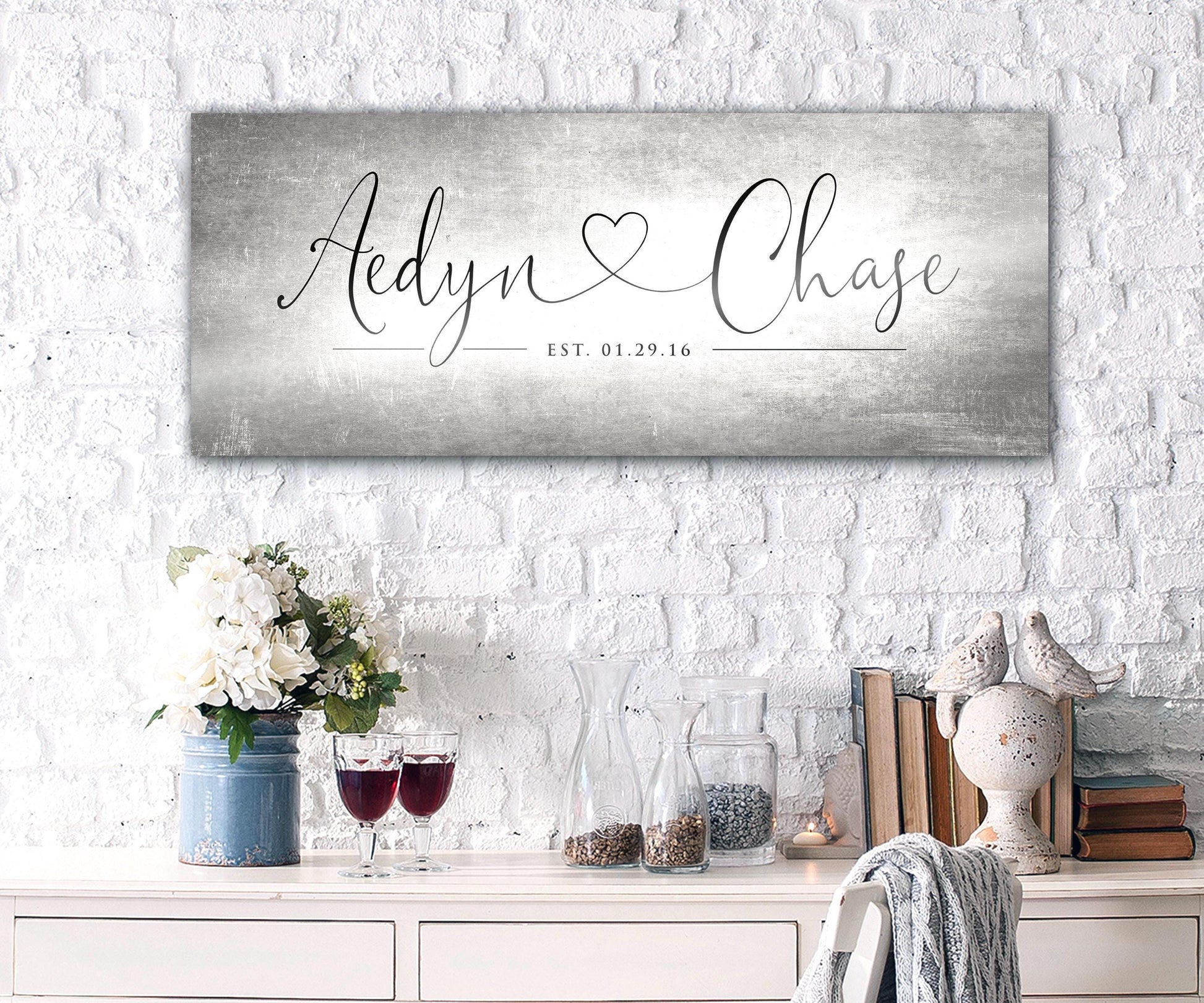 Unique Anniversary gifts like Honeycomb Proverbs' Personalized Heart Name signs are popular for ten year wedding anniversaries. One of their tarnished metal Heart Name sign hangs above an accent table. The sign is extra large sized and features personalized names connected by a heart in a whimsical calligraphy font. The couple's established date sits beneath their names to mark their special day.