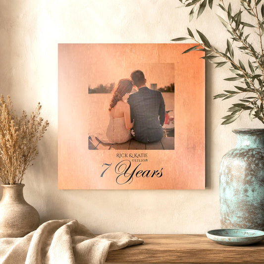 Beautiful Square Wall Decor on copper metal with "7 Year"s, and the names Nicole, and Robert, and the date 11.02.11"
A cropped portrait sits center, above the personalized inscription, and appears to also be in copper, with slight color tones showing through the copper base of the metal. The photo is of a bride and groom sitting together on a dock, shot from behind them with the water and mountains in the background. The artwork hangs on the wall of an elevated home space.
