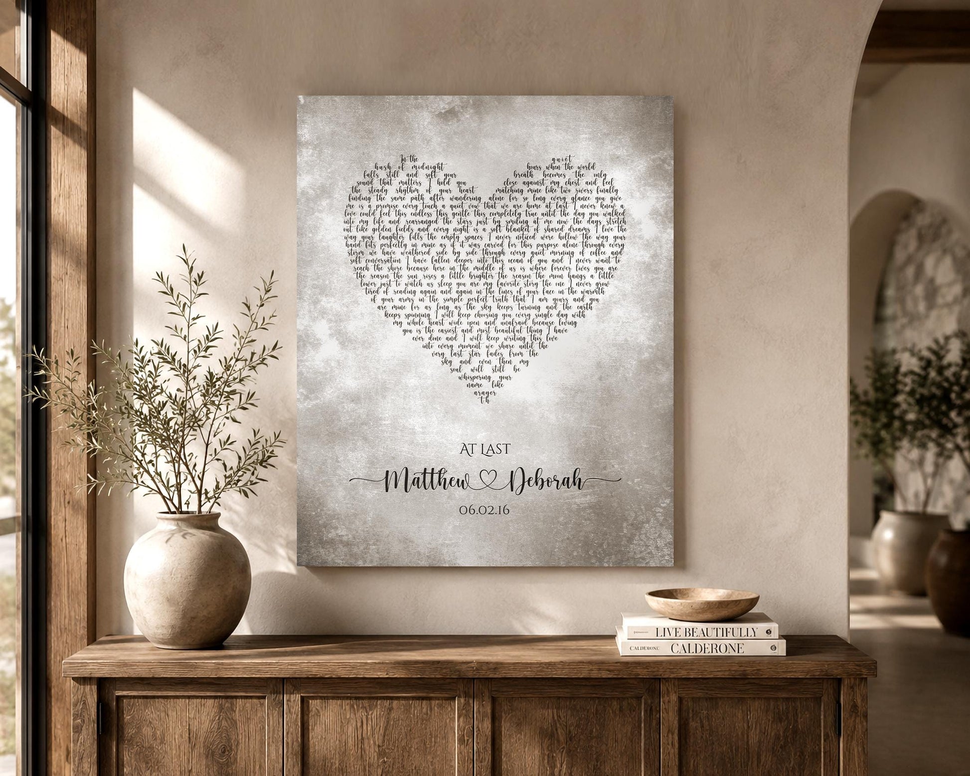 A tarnished metal artwork with custom lyrics shaped into a heart hanges on a wall near a window. The artwork is also personalized with the names of a married couple inscribed in a whimsical calligraphy font and joined by a decorative heart. Their union date sits beneath.  The song featured is At Last.