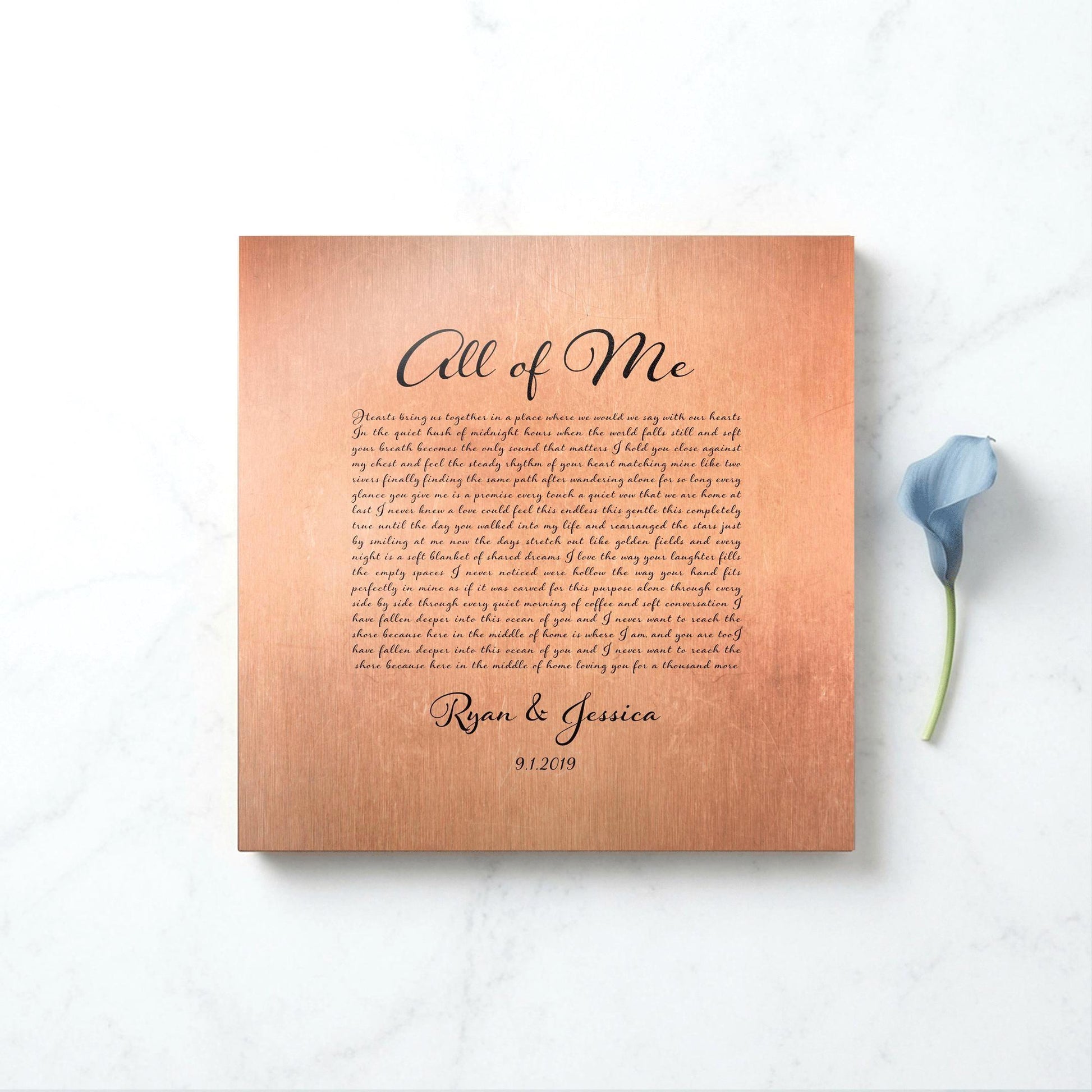 A copper metal song plaque sits on a marble surface with a blue lily accenting the right side of the frame. The song name is All of Me, and the plaque is finished with the names of a married couple and their union date, beneath a paragraph of song lyrics. The inscription is in black lettering in a calligraphic style letterhead.