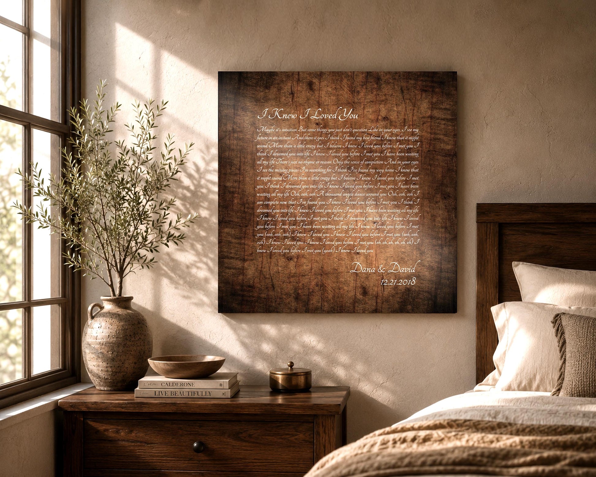 Wooden Song Lyrics Wall Art on a Stained birch square panel, with song lyrics inscribed with a calligraphy font. The song lyrics art is also personalized with the name of a married couple and their established date. It hangs on a wall in a rustic bedroom setting and sunlight comes through the window and landing on the wood artwork.