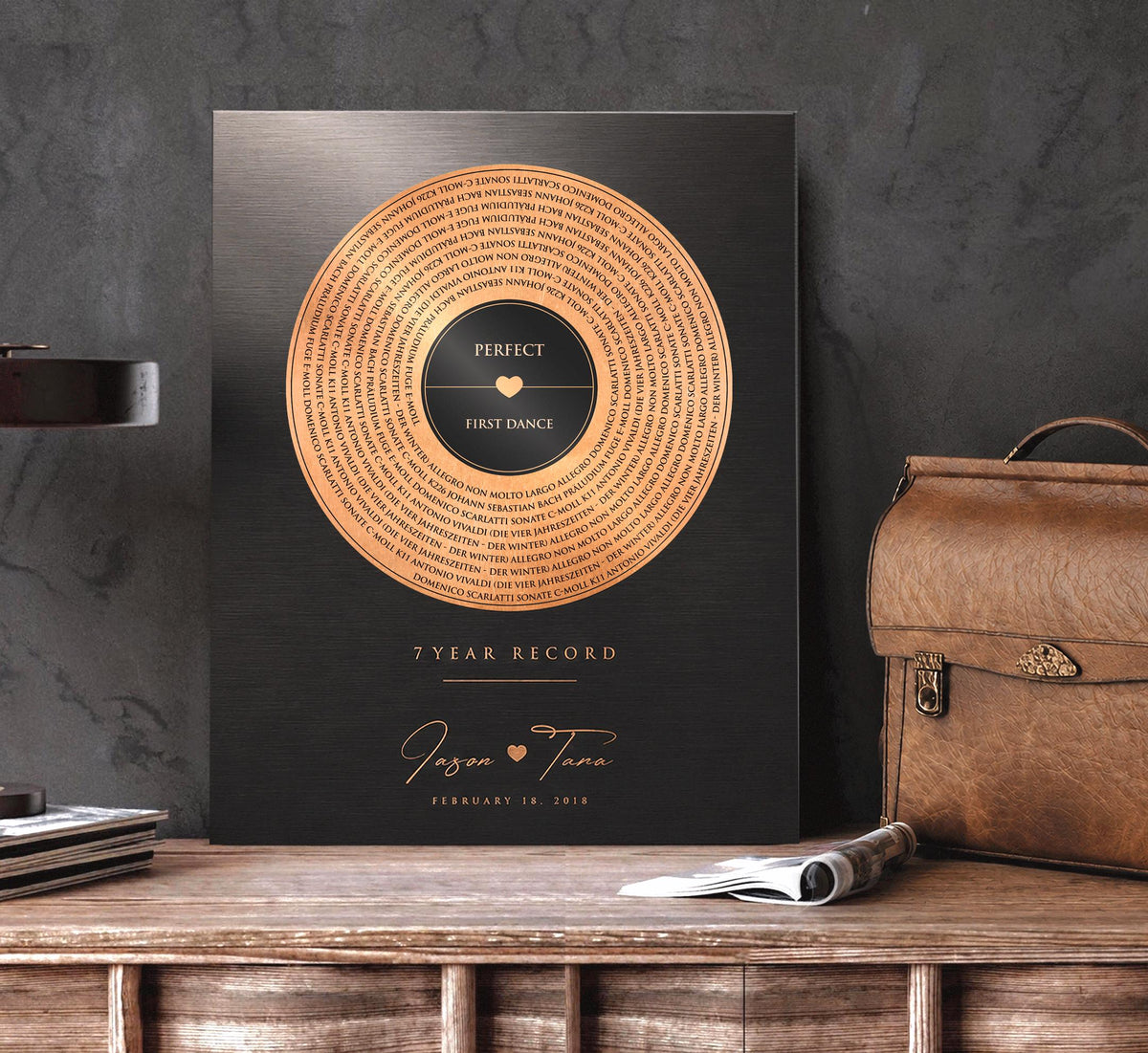 7 year record anniversary art or 22 year copper anniversary record sits on a tabletop next to a leather bag. Metal artwork features a vinyl record motif in distressed copper, with song lyrics over-laid. A heart joins the married couple's names beneath the proclamation of 7 or 22 years, and the personalization is completed with a union date to mark the official wedding day on the anniversary year tribute.