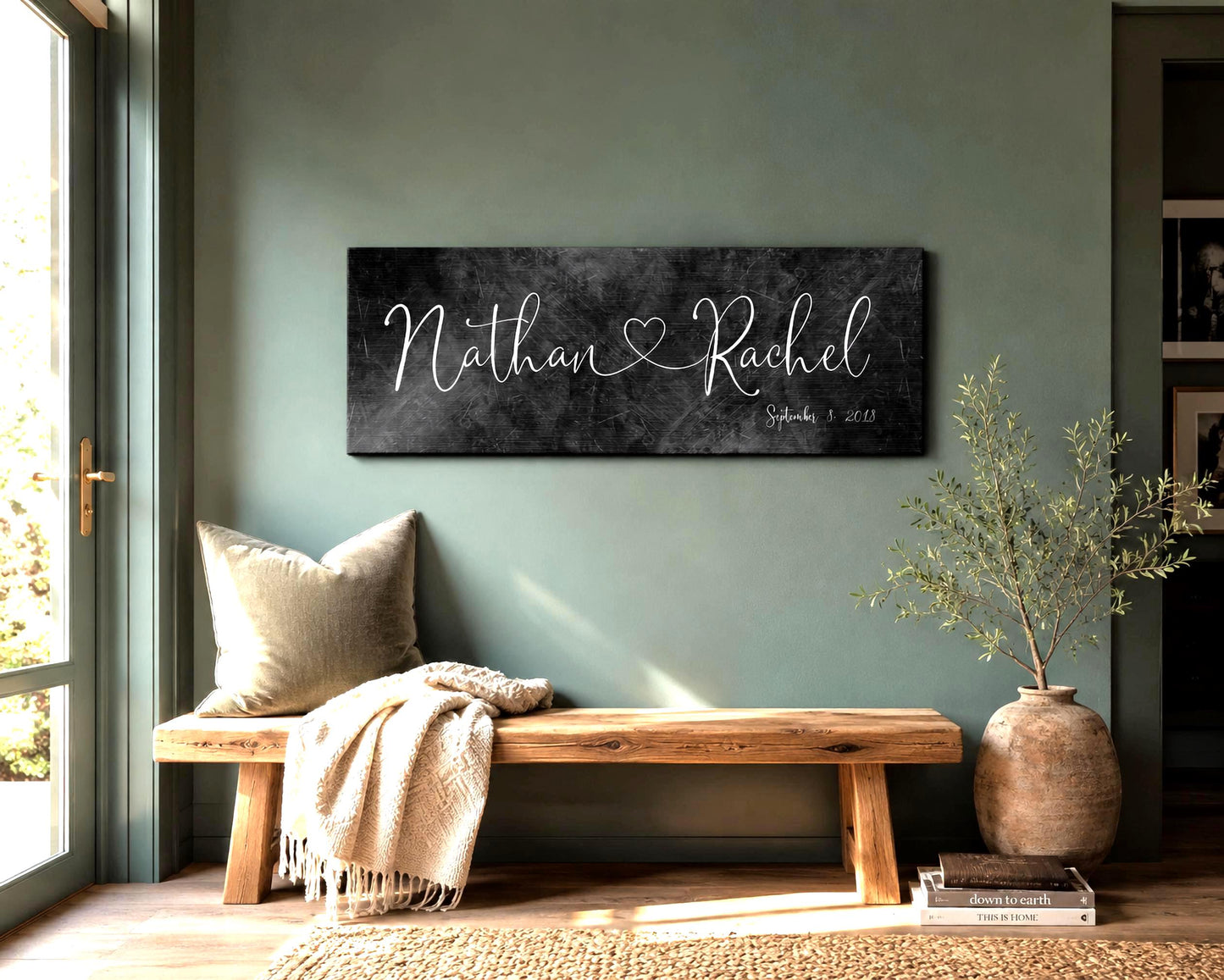 An wide format iron metal finished name sign with the names Nathan and Rachel in a calligraphy style lettering and connected by a whimsical heart. The Est. date is shown in the lower right corner - hangs on a muted green wall above a wooden bench with a throw and pillow. An indoor plant sits beside the bench and daylight falls across the scene from a window on the left side of the frame.