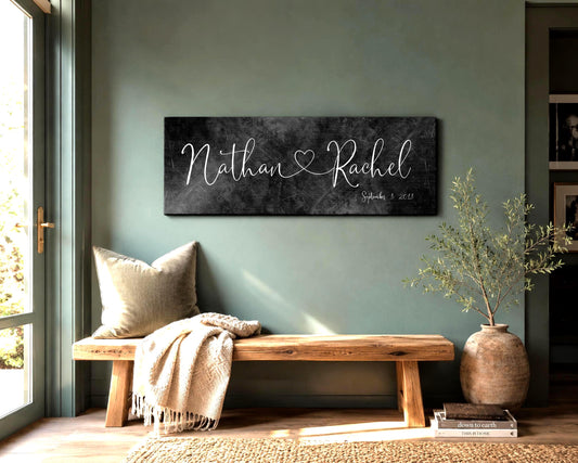An wide format iron metal finished name sign with the names Nathan and Rachel in a calligraphy style lettering and connected by a whimsical heart. The Est. date is shown in the lower right corner - hangs on a muted green wall above a wooden bench with a throw and pillow. An indoor plant sits beside the bench and daylight falls across the scene from a window on the left side of the frame.