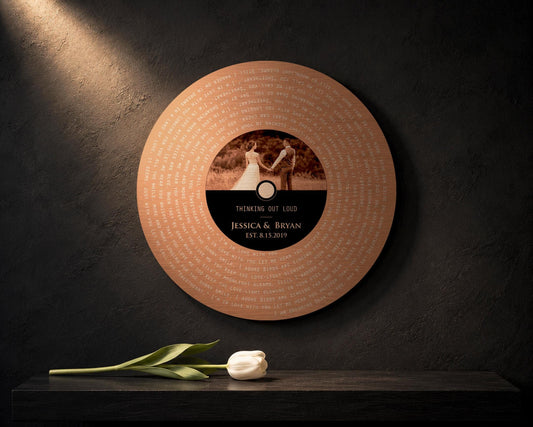 Round Copper Metal Vinyl Song Lyrics Photo Record:
Round photo plaque shaped and designed like a vinyl record. Record personalized with the customer's photo, and favorite song with a date included. A white tulip sits on the shelf below the round photo gift which is mounted onto a charcoal wall.