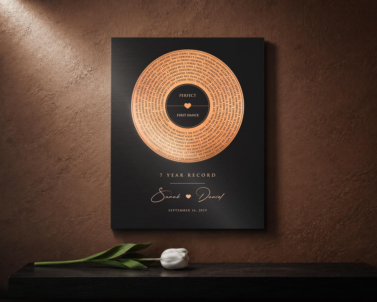 Copper anniversary gift is mounted against copper toned wall and is a black metal wall plaque or sign which is a 7 year anniversary song record, personalized with the names and wedding date of a couple celebrating their 7th wedding anniversary, and the song lyrics from their first dance circling around the tarnished copper record motif.