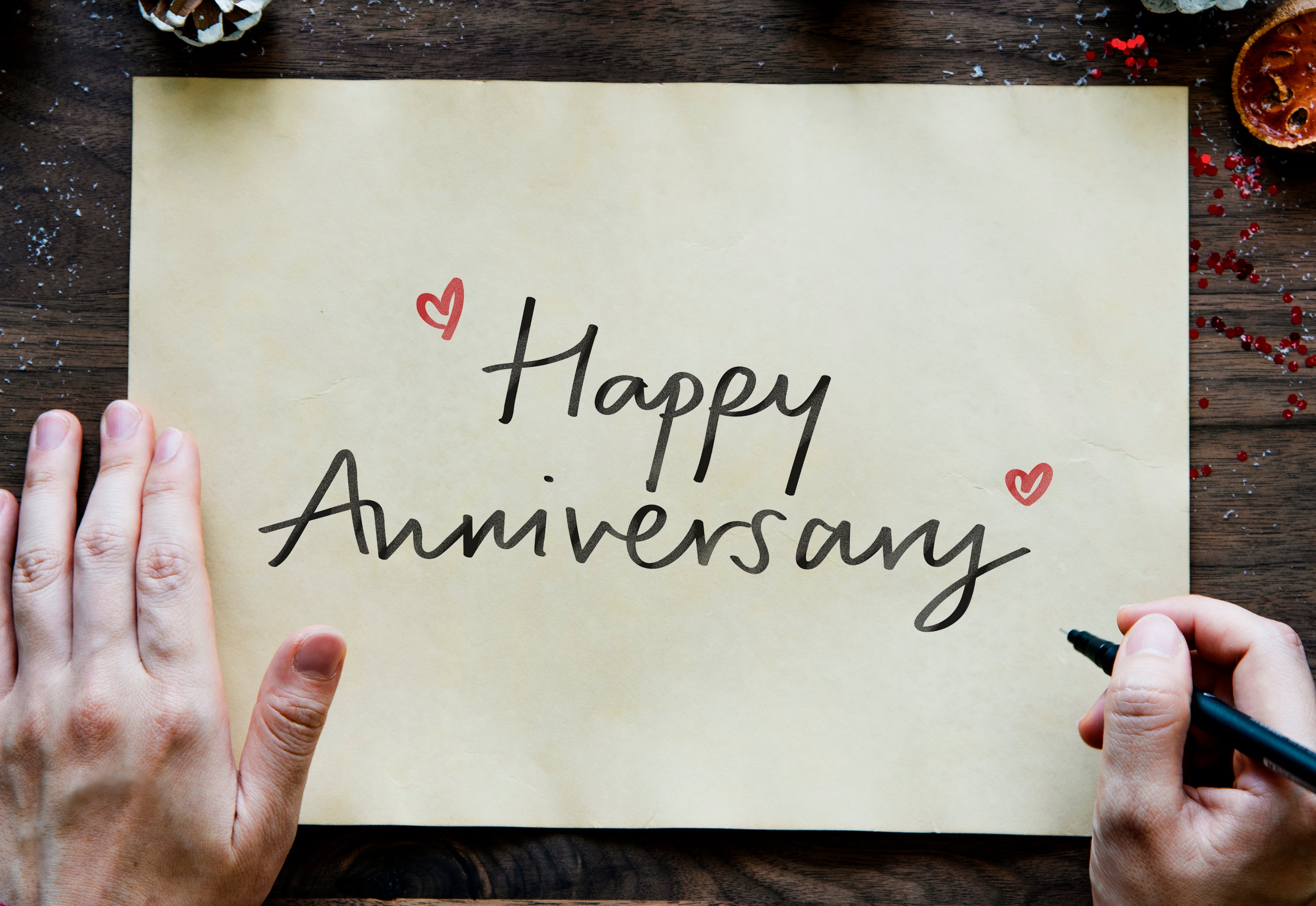 Handwritten 'Happy Anniversary' on a piece of paper with hearts, held by two hands on a wooden surface.